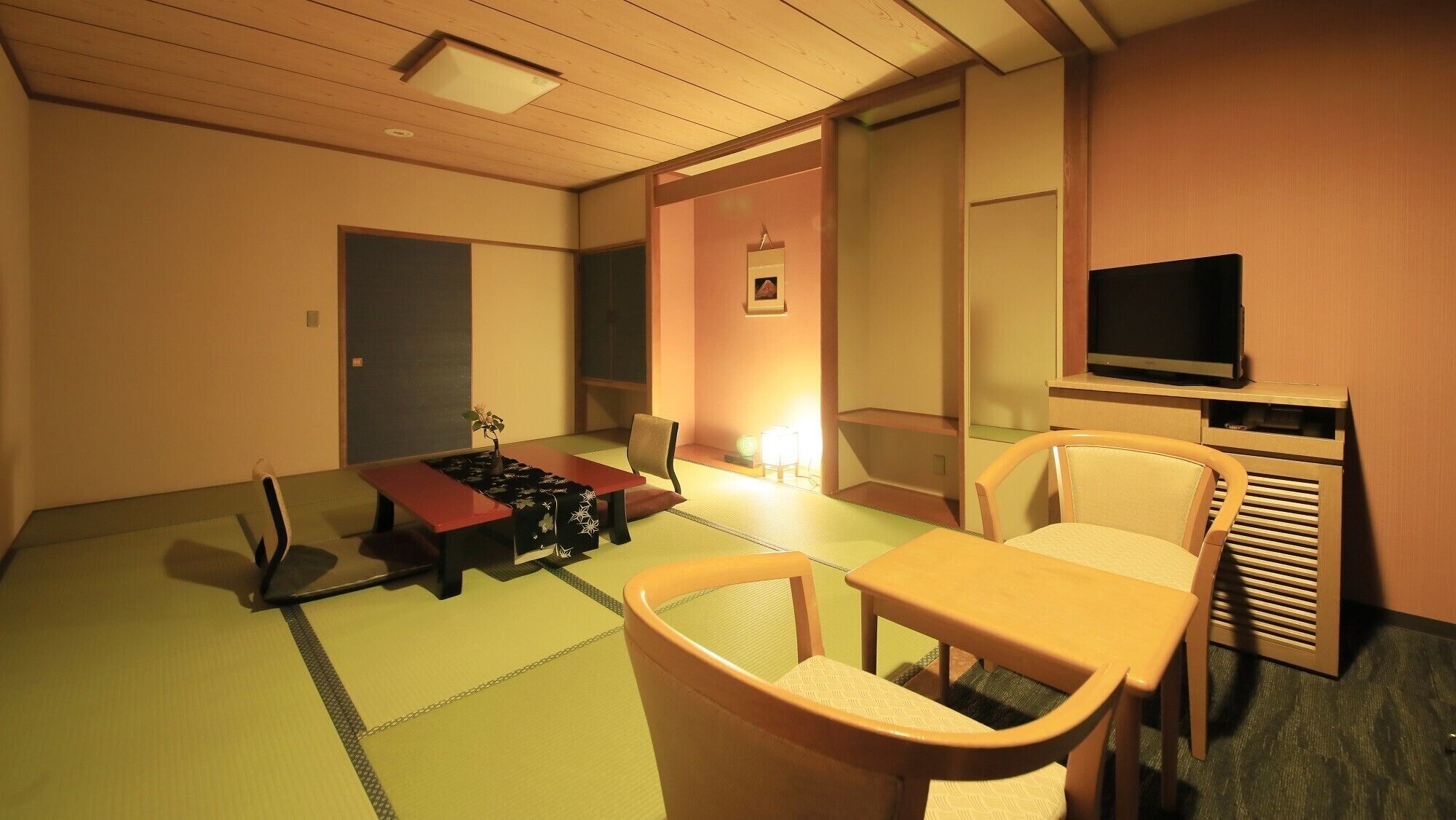 Une pièce avec un sol recouvert de tatamis, un futon servant de lit, une télévision et une petite table avec des chaises.