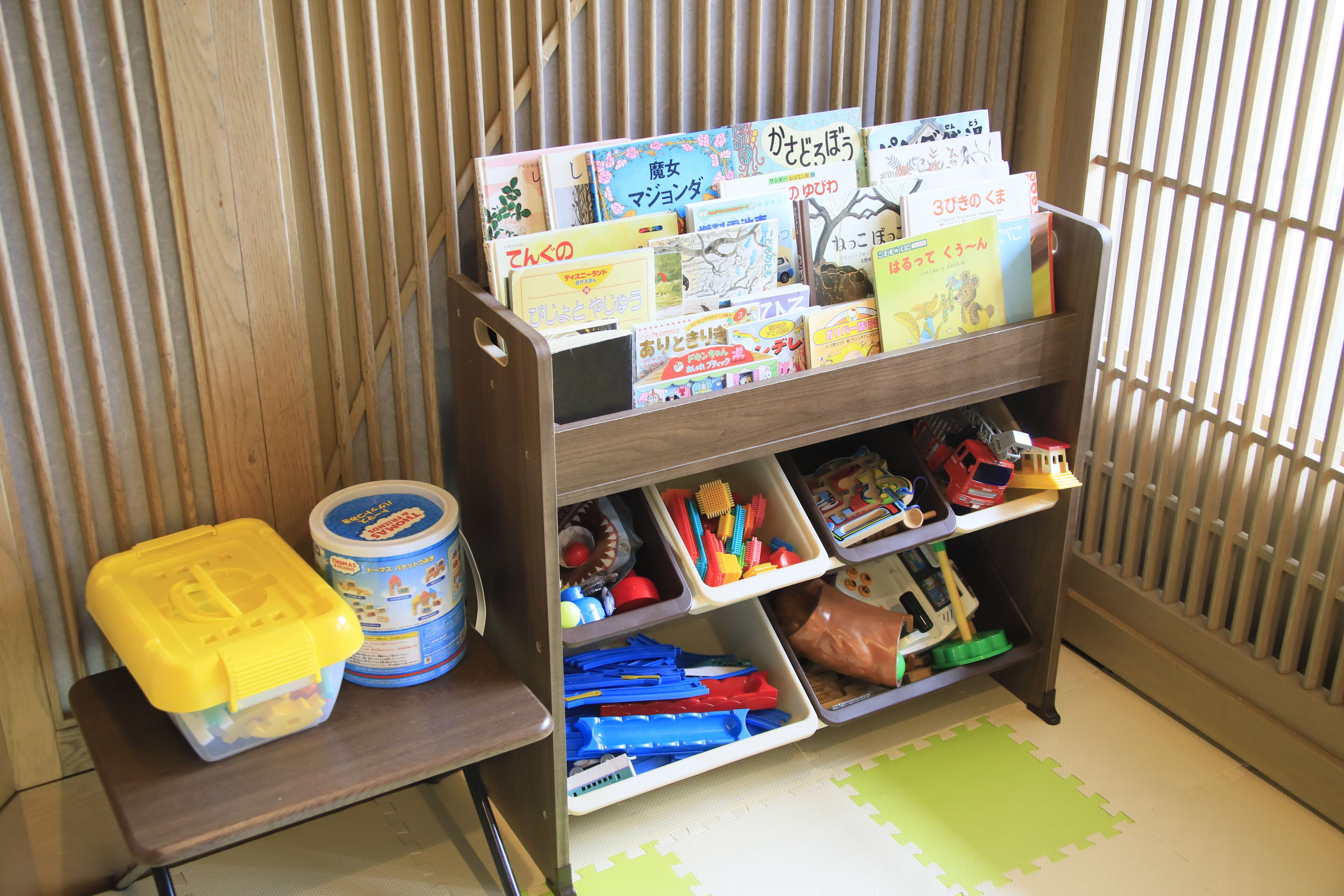 Une étagère en bois avec des livres et des jouets pour enfants, un récipient en plastique et un autre récipient bleu doté d’un couvercle.