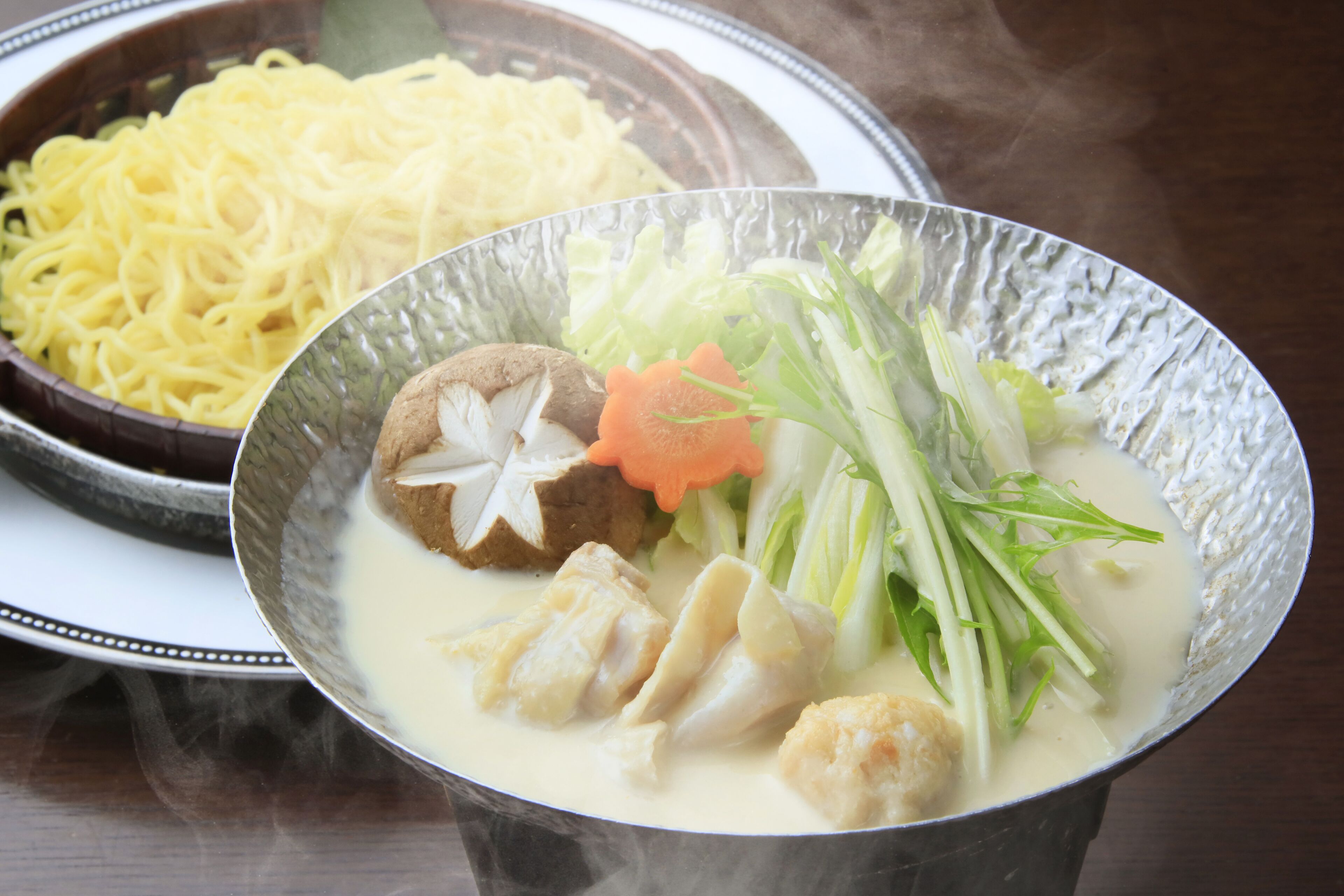 A bowl of soup with vegetables and meat, and a plate of noodles in the background.