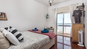 Quarto com cama, almofadas, mesa de cabeceira, luminária pendente, janela com cortinas e vista para o mar, além de um ventilador.