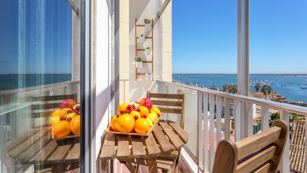 Varanda com mesa e cadeiras de madeira, bandeja de frutas e vista para o mar e barcos.