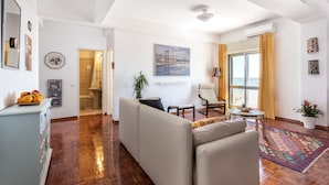 Uma sala de estar ampla com sofá, poltrona e uma mesinha. Há uma janela grande com cortinas, um quadro na parede e um vaso de planta.