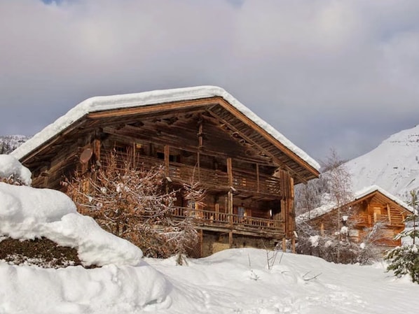 Um chalé de madeira com telhado coberto de neve, rodeado por árvores e montanhas cobertas de neve.