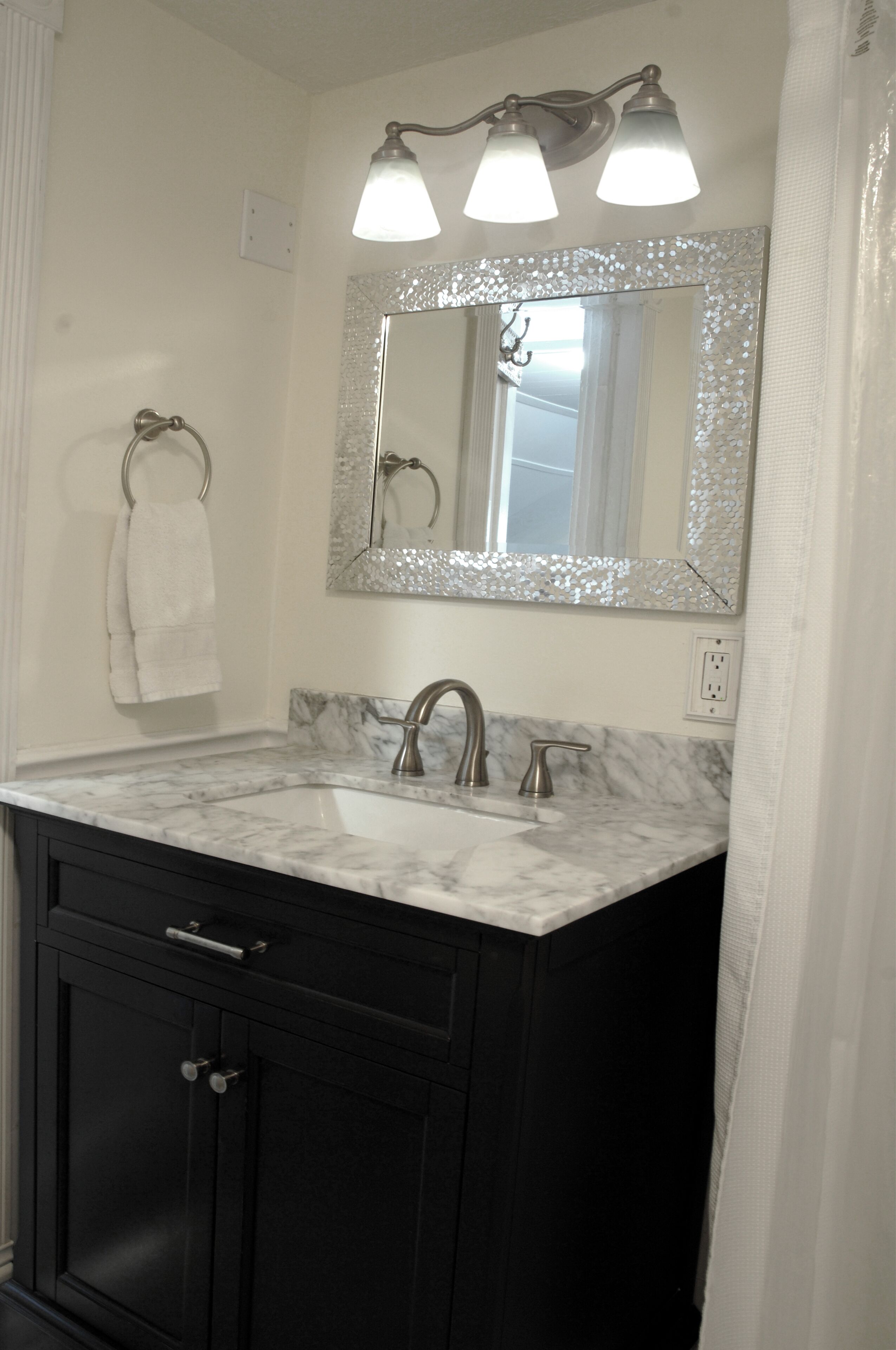 Un baño con lavamanos de mármol, armarios negros, un sujetarrepuestos para toallas y un espejo con marco decorativo.