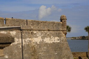 A weathered stone wall with a chain link barrier, overlooking a body of water with a distant view of a marina and buildings. - New! Victorian Apartment. Park & Walk Everywhere in Town (St. Augustine)