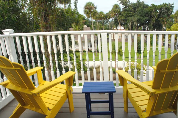 A balcony with yellow Adirondack chairs and a blue table, overlooking a well-maintained lawn and a white fence. - New! Victorian Apartment. Park & Walk Everywhere in Town (St. Augustine)
