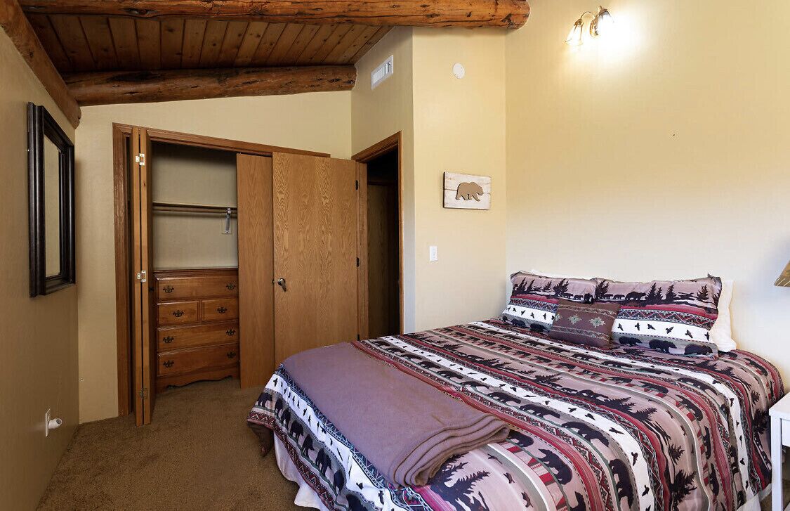 A bedroom with a wooden ceiling, a bed with patterned bedding, a wooden wardrobe, and a wall-mounted picture of a bear.