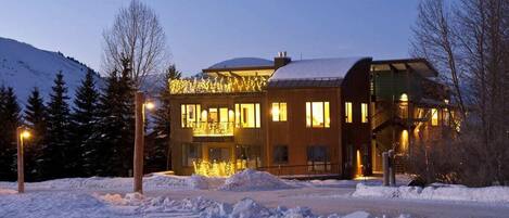 A modern building with a snow-covered roof, large windows, and a balcony with lights.