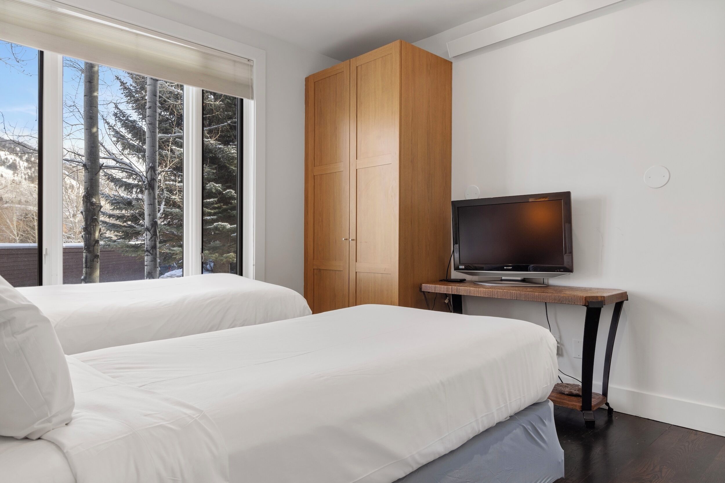 A hotel room with two beds, a TV on a small table, and a large window with a view of trees.