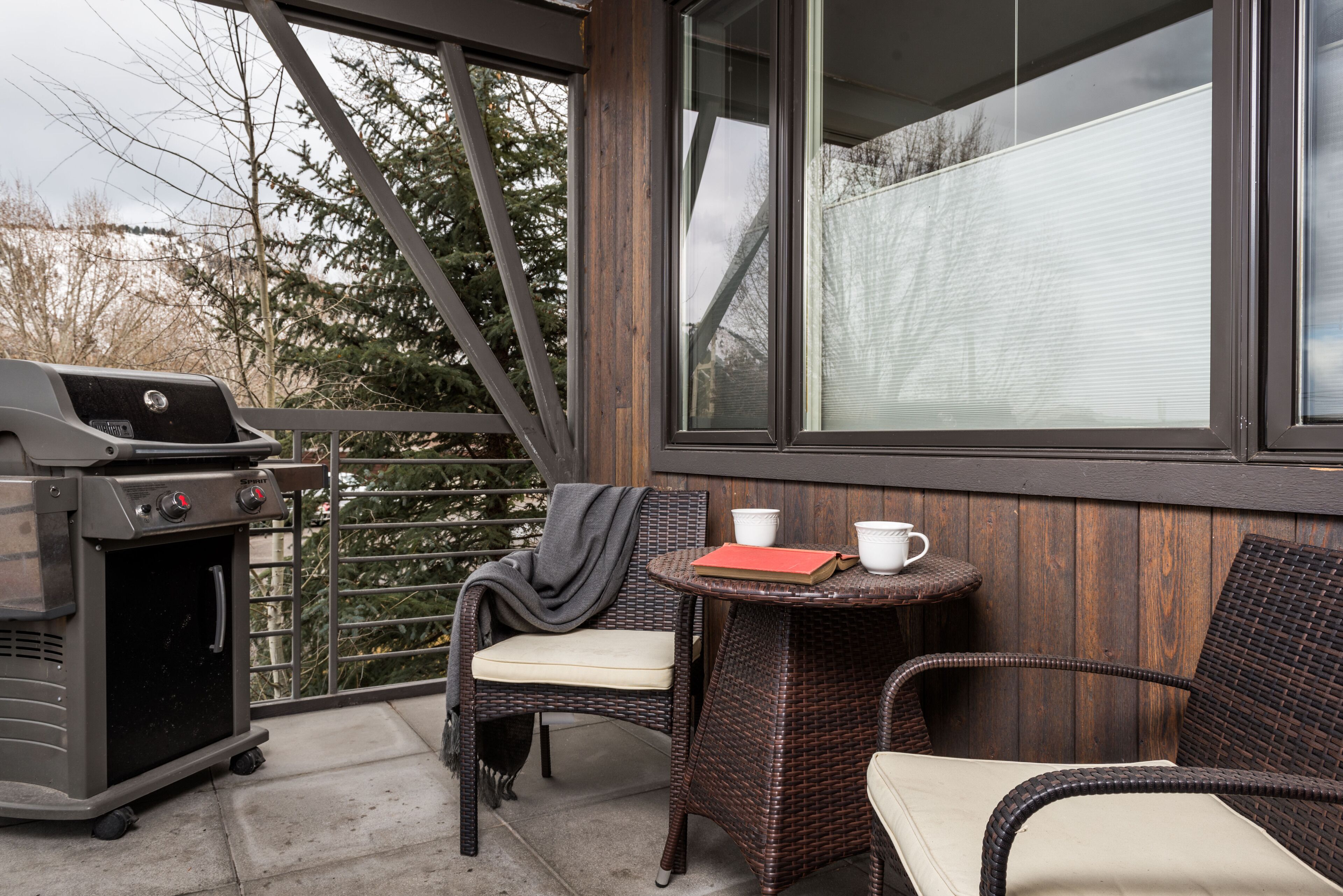 Een balkon met rieten stoelen, een klein tafeltje met kopjes en een barbecue.