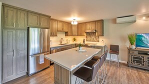 A modern kitchen with wooden cabinets, stainless steel appliances, a central island with bar stools, and a large window providing natural light.