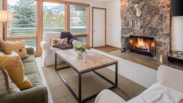 A cozy living room with a stone fireplace, a sofa, a coffee table, and large windows offering a view of trees and mountains.