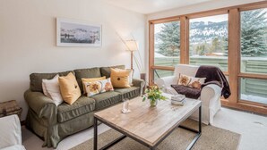 A living room with a green sofa, a white armchair, and a wooden coffee table. There is a window with a view of a snowy landscape.