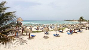 A beach with thatched umbrellas and lounge chairs, a palm tree, and a small structure on the left.