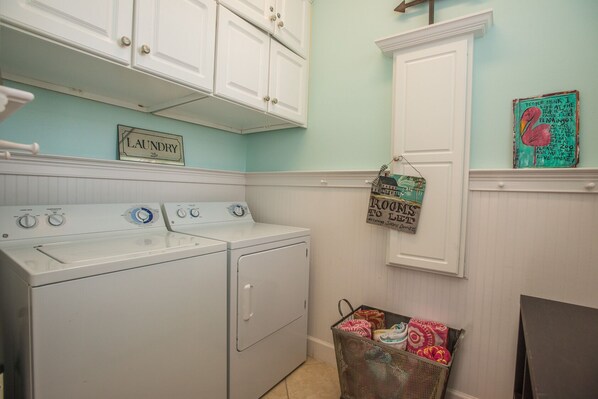A laundry room with a washer and dryer, a basket of folded clothes, and a sign indicating rooms to let. - Spacious Gulf View, 110 steps to the gulf, elevator, beach gear included! (Port St Joe)