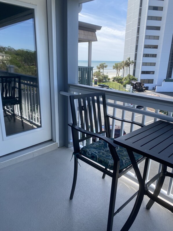 A balcony with a table and chairs, overlooking a beach and a parking area.