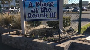 A sign for 'A Place at the Beach III' with a view of a parking lot and buildings in the background. - APATBIII Ocean Side/Ocean View 9520 SHORE DR 3P Family friendly.  (Myrtle Beach)