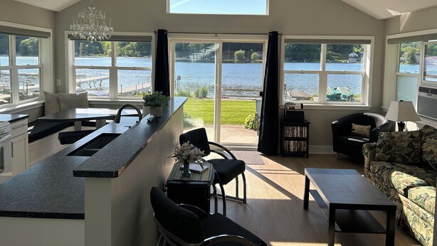A spacious kitchen with a large island, a dining area with chairs, a sofa, and a view of a lake through large windows.