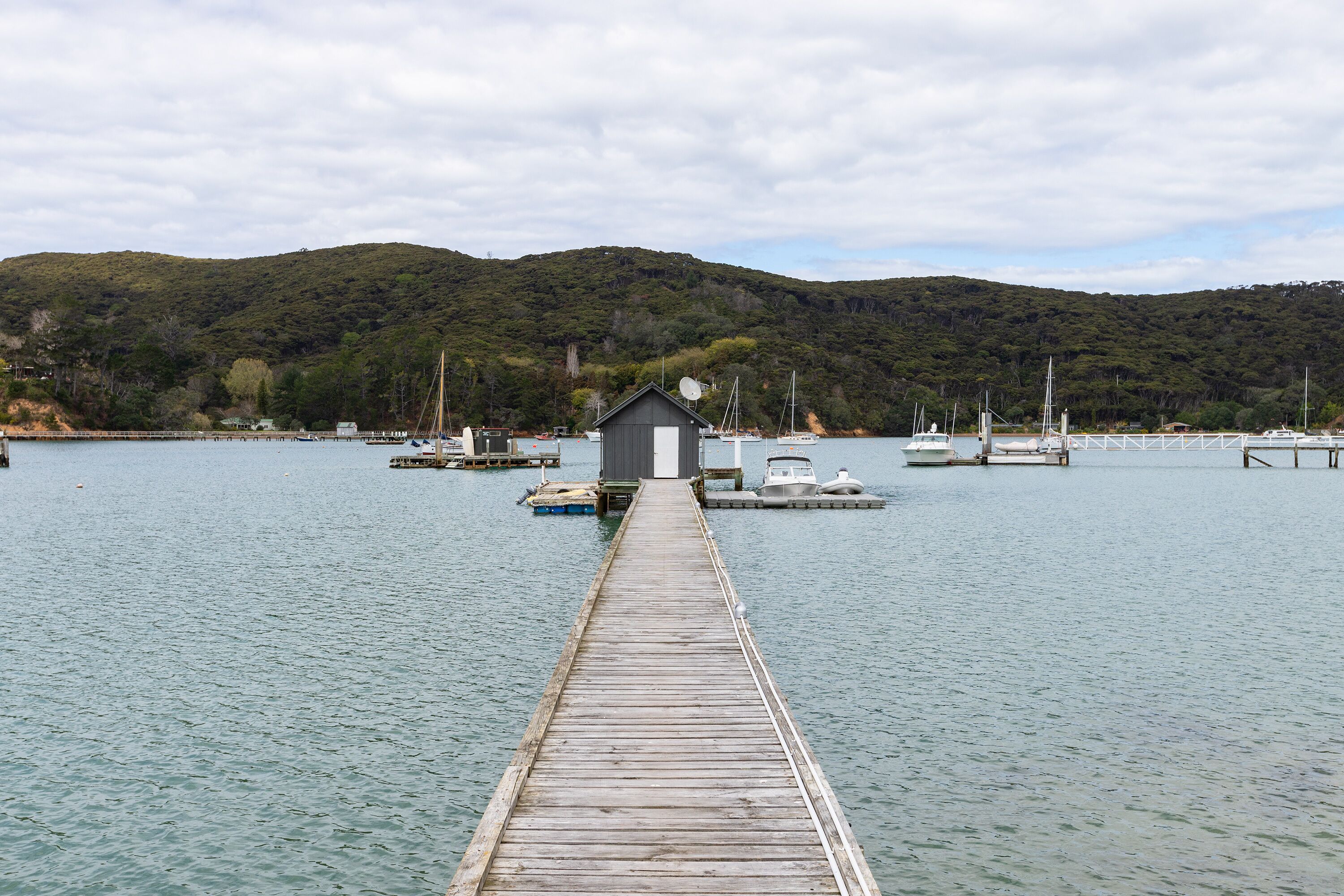 A wooden pier leading to a small dock with a house-like structure at the end. Boats are docked nearby, and hills are visible in the background.