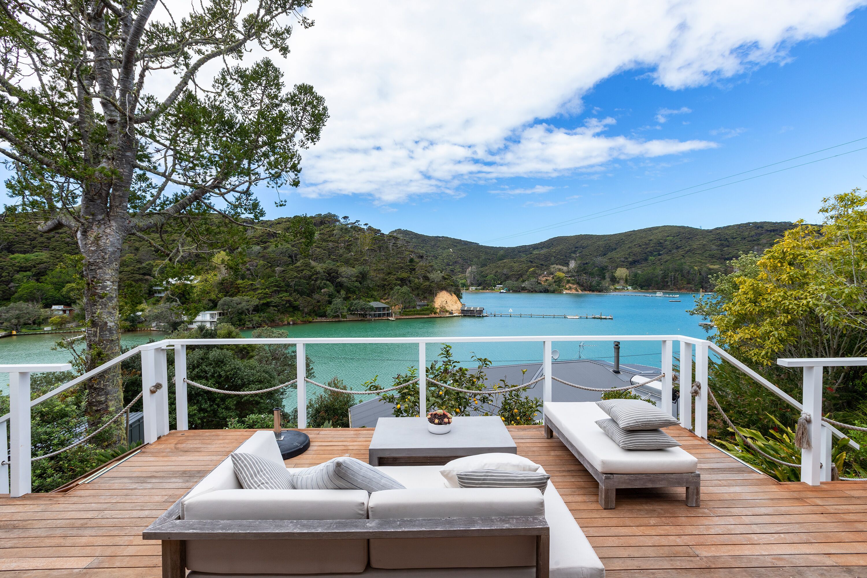 A wooden deck with outdoor seating, overlooking a turquoise bay and a forested hillside.