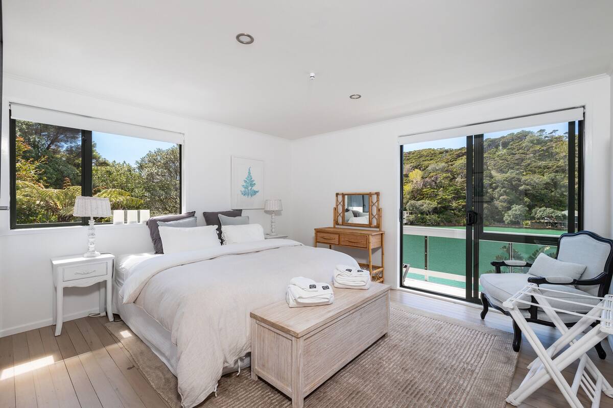 A bedroom with a large bed, a wooden bench with towels, a nightstand, a dresser, a chair, and a balcony with a view of trees.