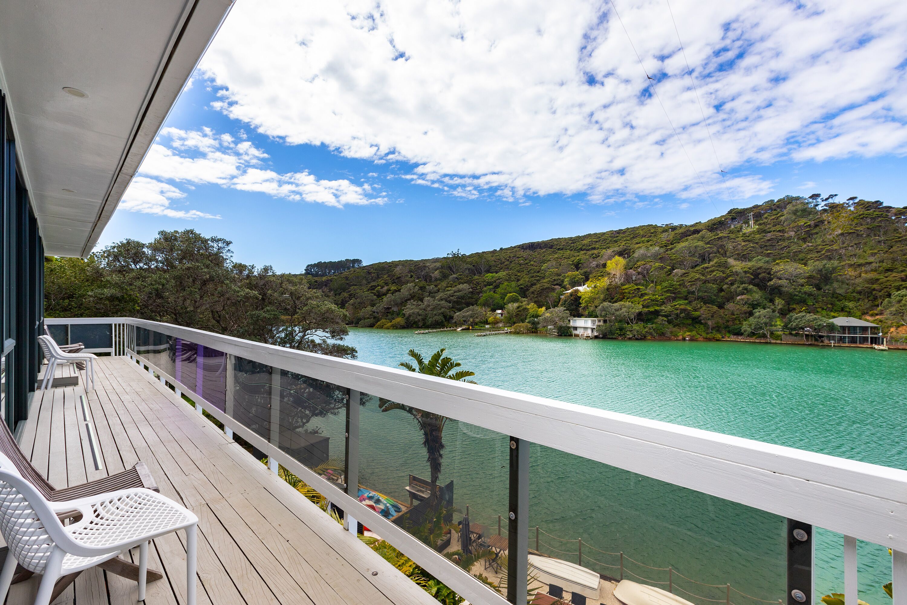 Terraza de madera con sillas blancas y baranda de cristal con vistas a un lago y una ladera boscosa.
