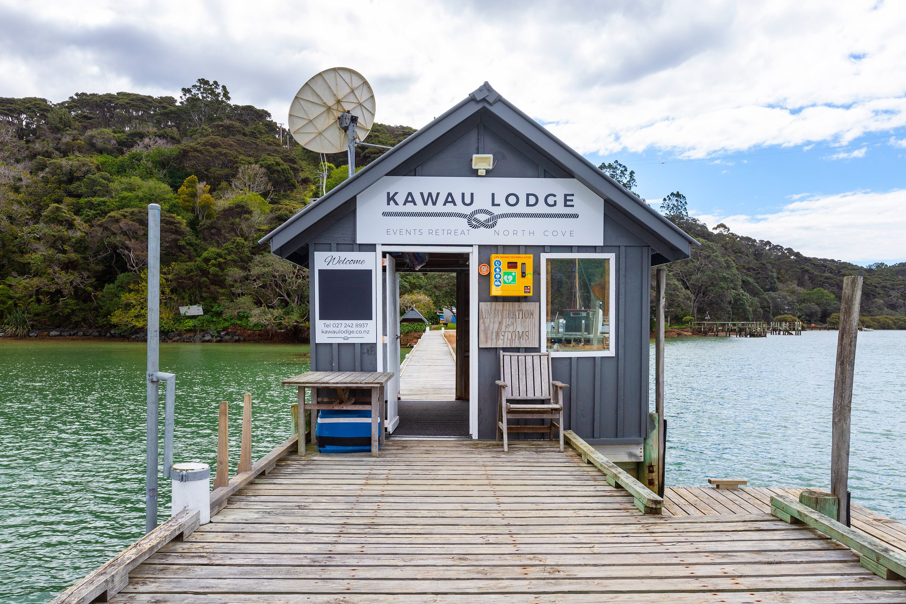 A wooden pier leading to a lodge with a sign reading "Kawau Lodge Events Retreat North Cove."