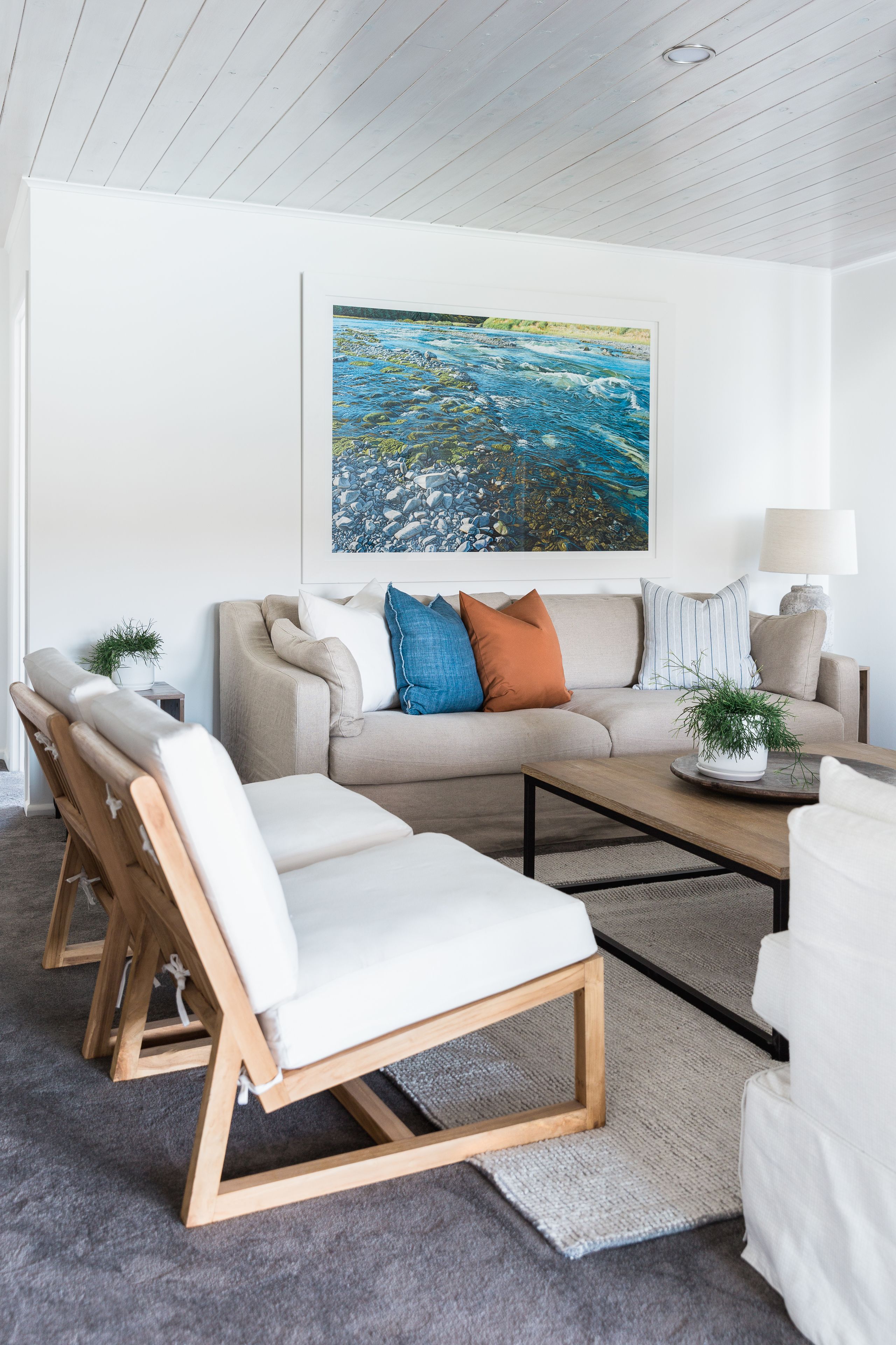 A modern living room with a beige sofa, two white armchairs, a coffee table, and a painting of a river landscape on the wall.