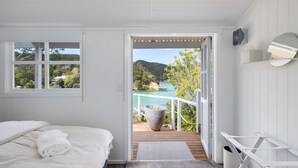 A bedroom with a bed, a window, a door leading to a balcony, and a view of a lake and mountains. - Kawau Lodge Boutique Resort (Kawau Island)