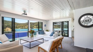 A modern living room with a large window overlooking a scenic view, a white sofa, a wooden chair, and a black-framed clock on the wall. - Kawau Lodge Boutique Resort (Kawau Island)
