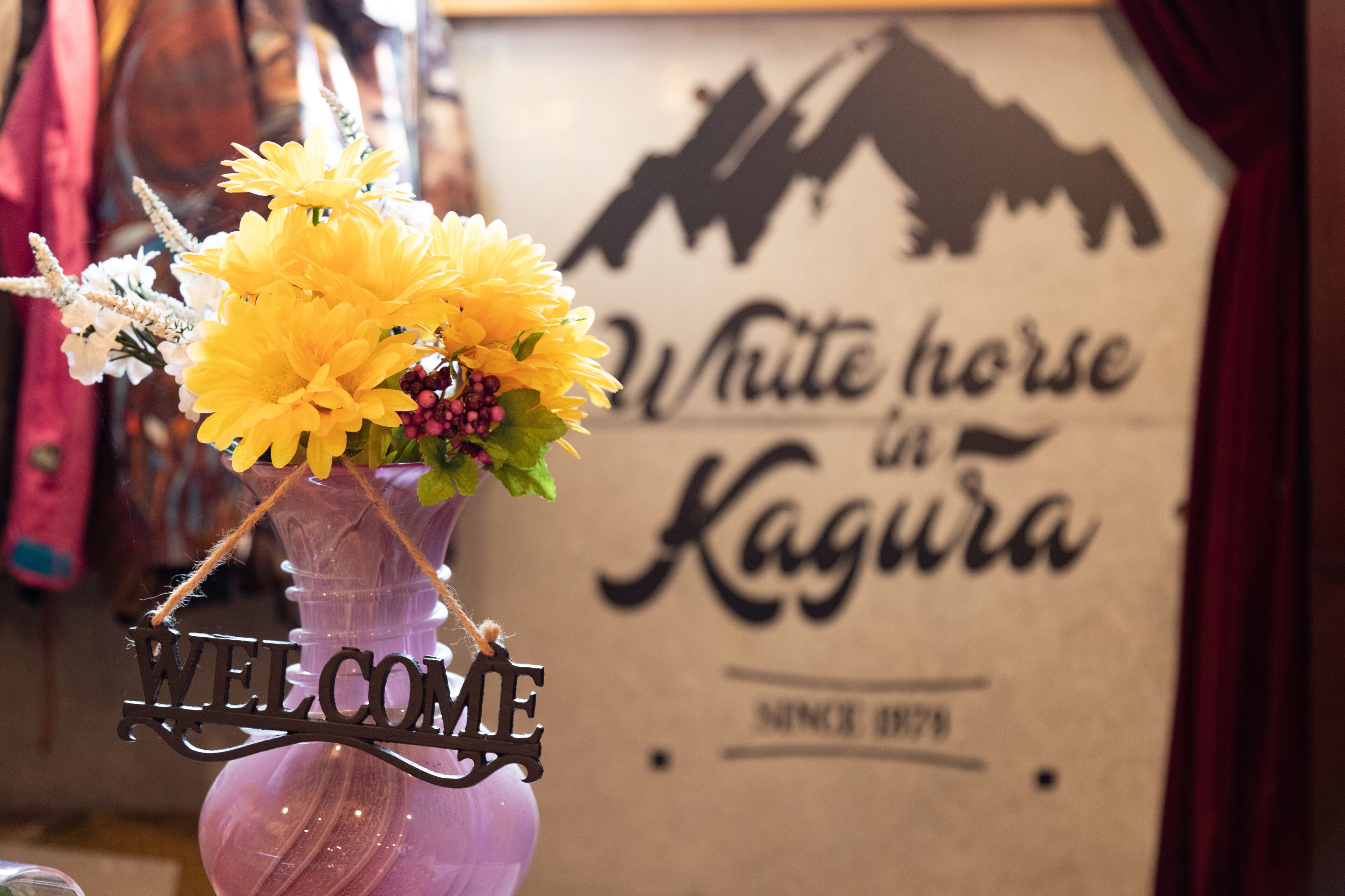 A purple vase with yellow flowers and a 'WELCOME' sign. In the background, there's a sign with 'White Horse in Kagura' and 'Since 1879'.