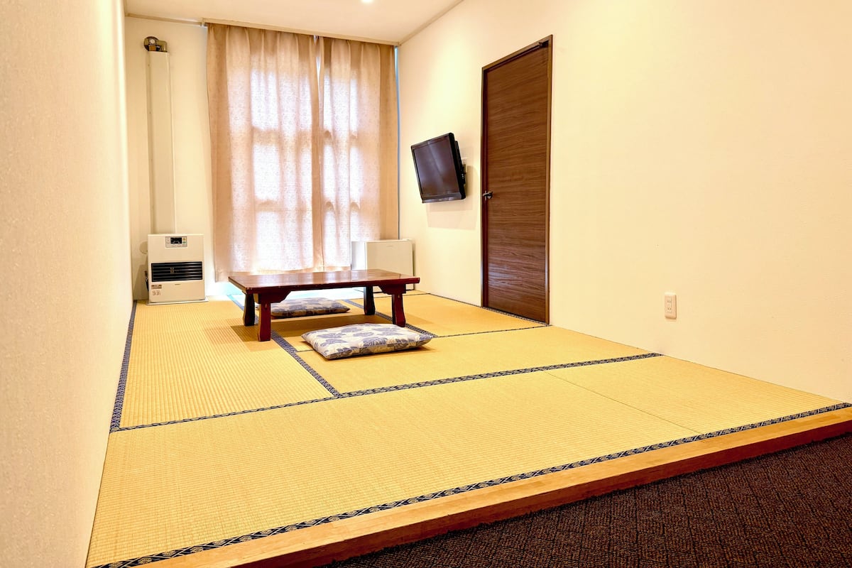 Una habitación con tatami, una mesa baja y cojines, un televisor en la pared y una puerta corredera.