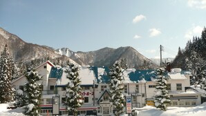 Un hotel con techo nevado y numerosas ventanas, rodeado de árboles cubiertos de nieve y montañas al fondo.
