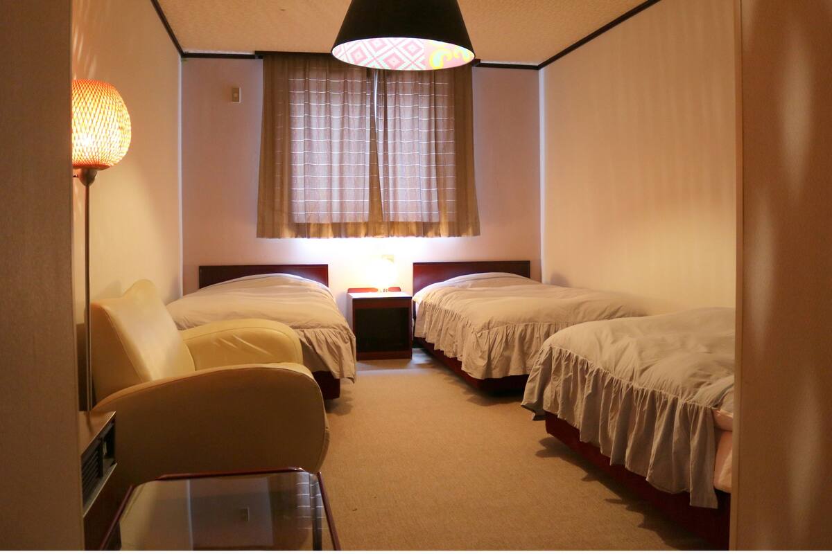 Habitación de hotel con dos camas individuales, una mesita, una silla y una lámpara.