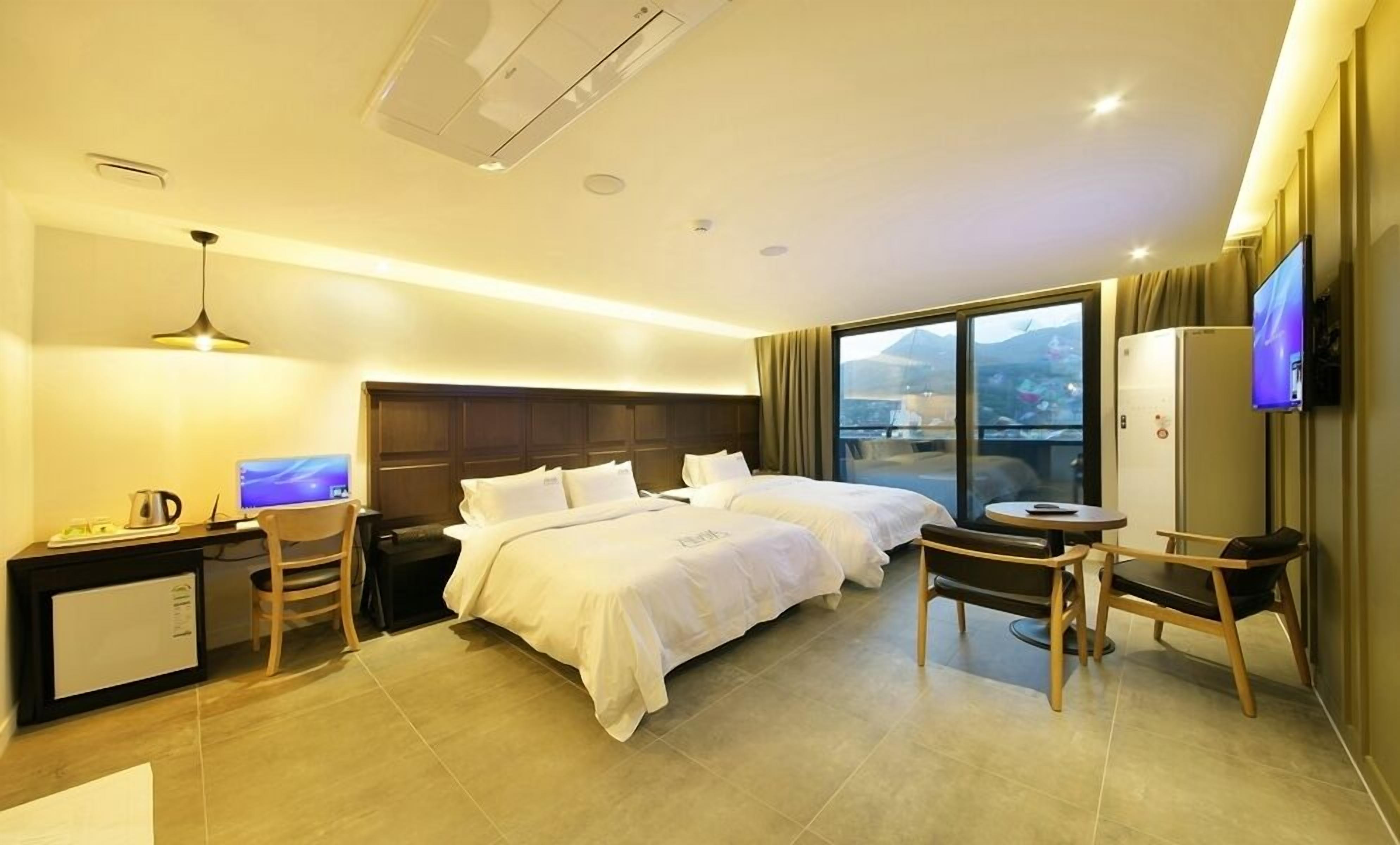 A hotel room with two beds, a desk with a computer, a chair, a TV, and a large window with a view.