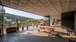Een terras buiten met een houten picknicktafel en banken, plantenbakken en uitzicht op een heuvel met gebouwen.