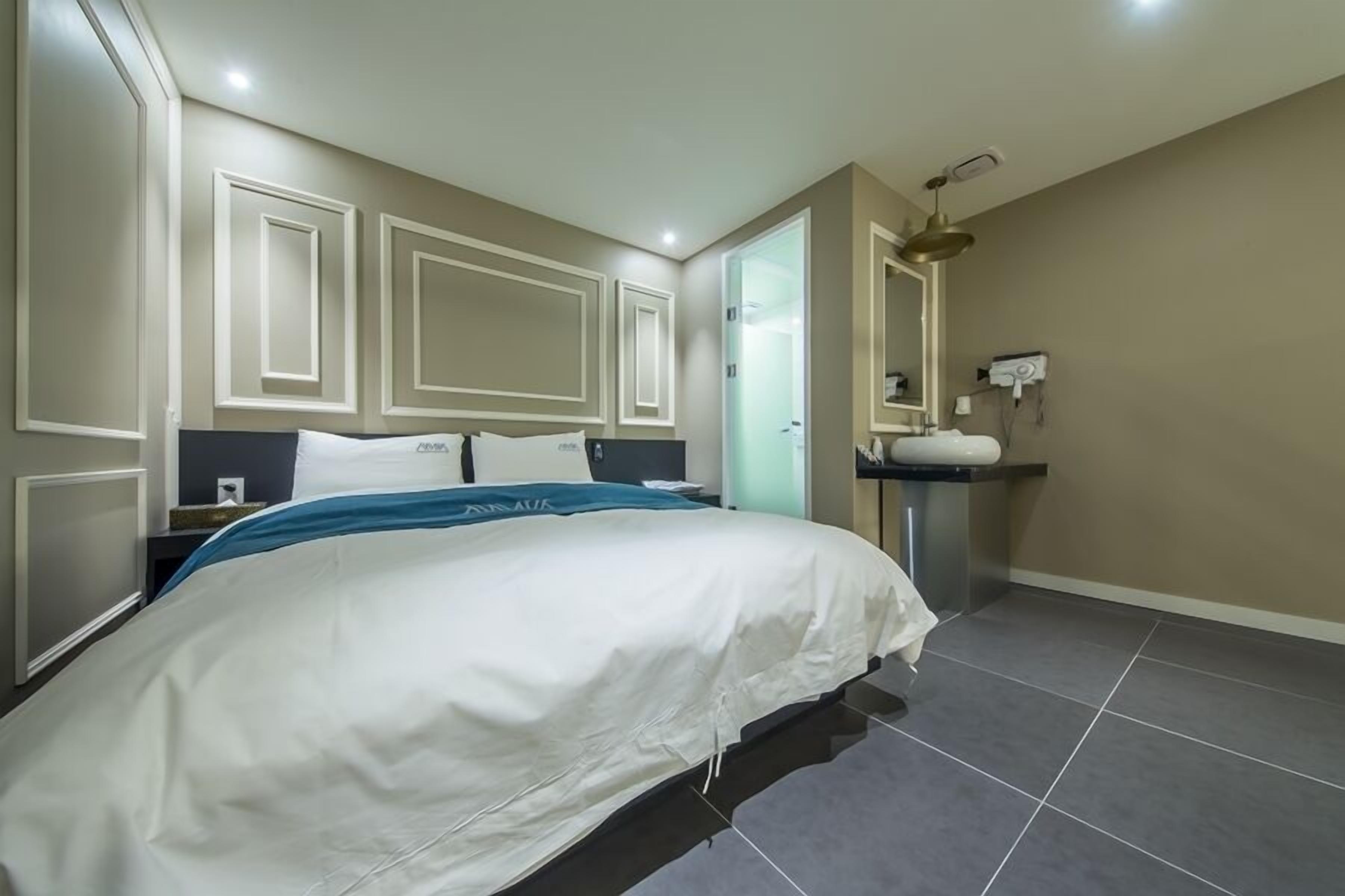 A hotel room with a large bed, a bedside table, a mirror, a sink, and a door leading to another room.