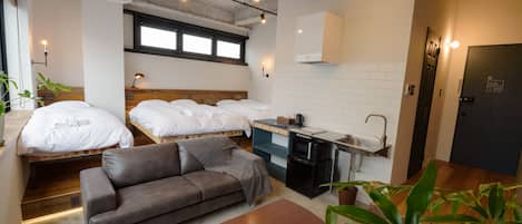 Una habitación de hotel compacta con dos camas individuales, un sofá, una kitchenette pequeña y un baño.