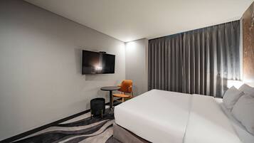 A hotel room with a large bed, a small table with a chair, a television mounted on the wall, and a window with curtains.
