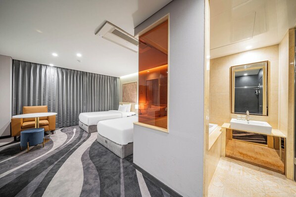 A hotel room with a bed, a small table with a chair, a large window, and a bathroom with a sink and mirror.