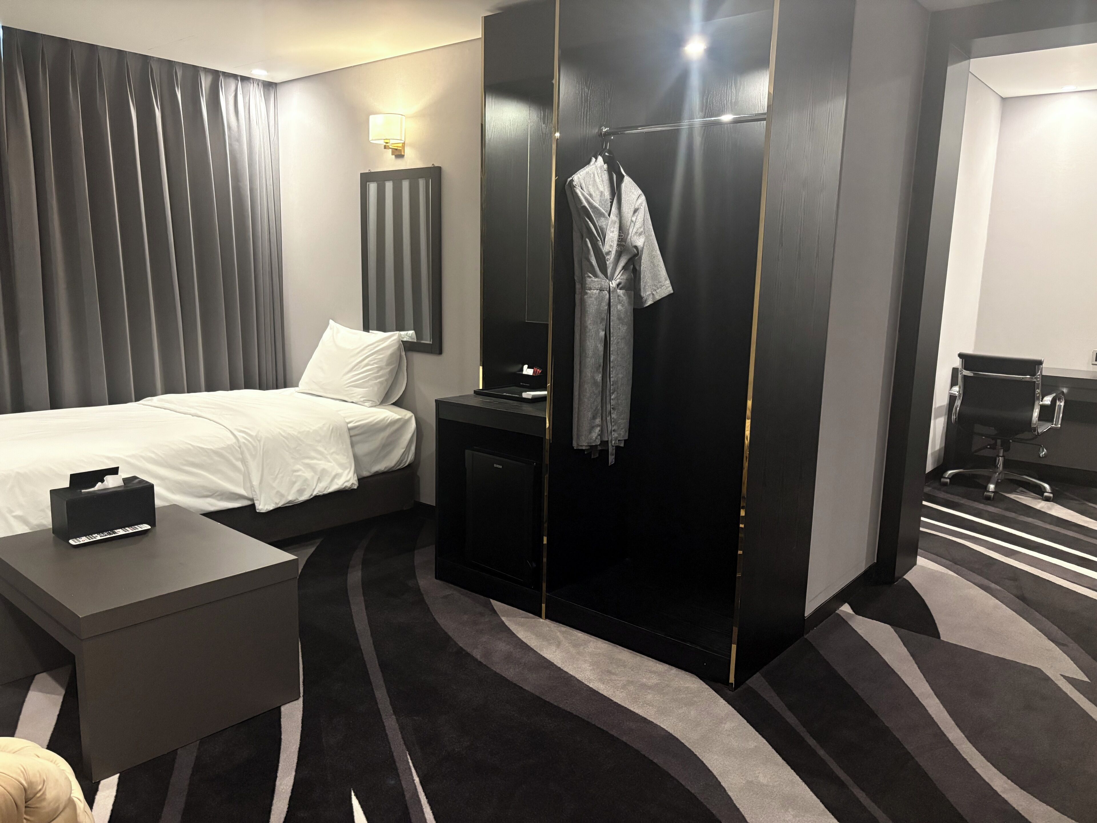 A hotel room with a bed, a desk, a robe hanging on a door, and a chair.