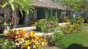 Ein Haus mit einem Strohdach und einer Veranda, umgeben von üppigen tropischen Pflanzen und einem gepflegten Garten.