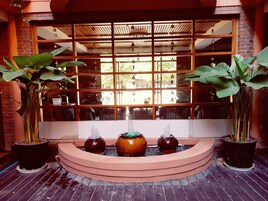 Una fuente de tres niveles, rodeada de plantas en macetas, frente a una ventana con diseño de celosía.