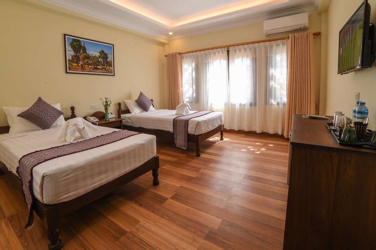 A hotel room with two beds, wooden flooring, a desk with a water bottle, and a painting on the wall.