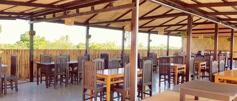 Une terrasse extérieure couverte, dotée de plusieurs tables et chaises, d’une clôture en bois et offrant une vue dégagée sur la végétation environnante.