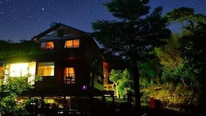 Une maison en bois à deux étages, dotée d’un balcon, entourée d’arbres et baignée par la lumière d’un ciel étoilé.