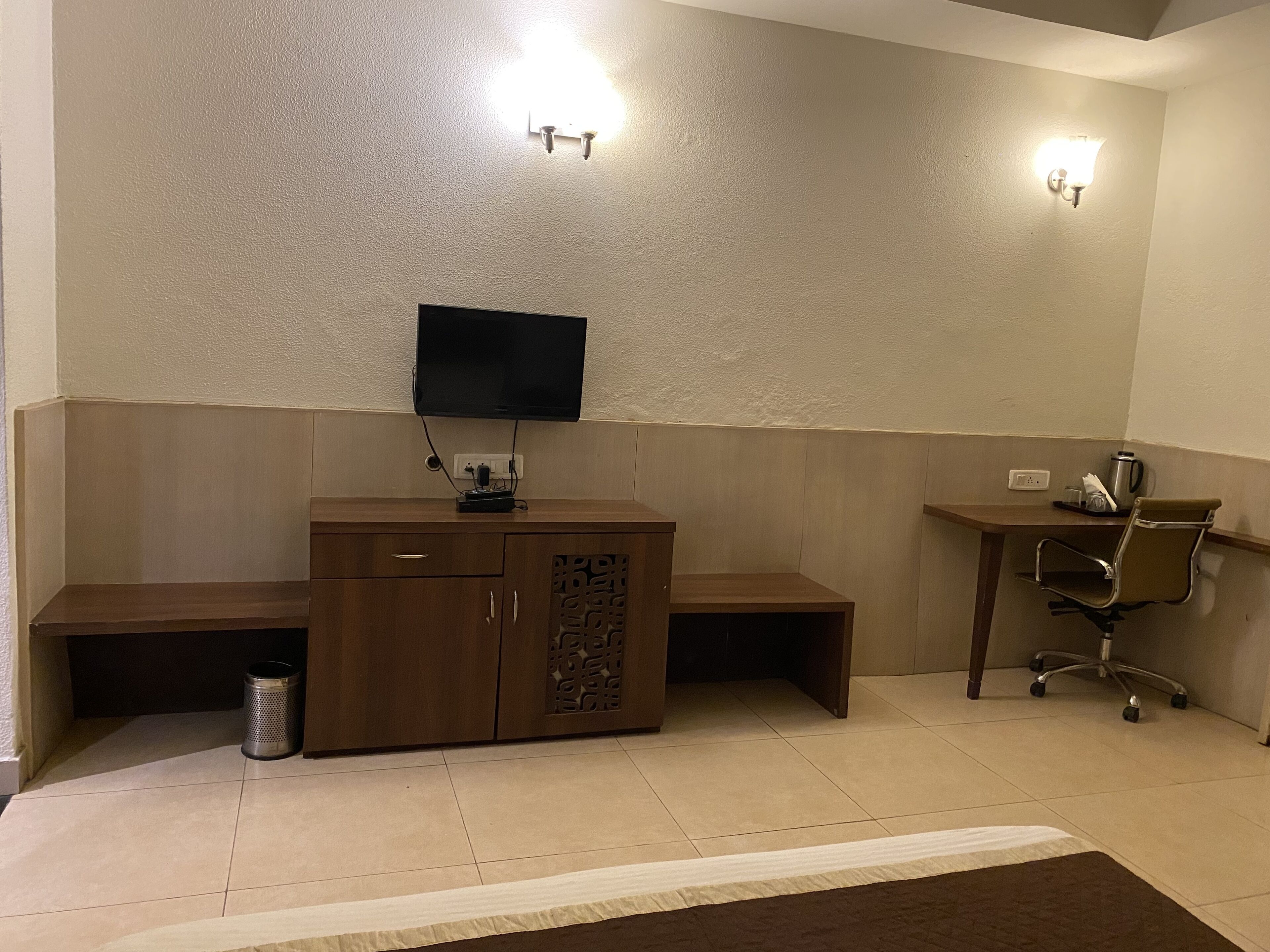 A small, well-lit room with a wooden desk, a chair, a television mounted on the wall, and a trash can.