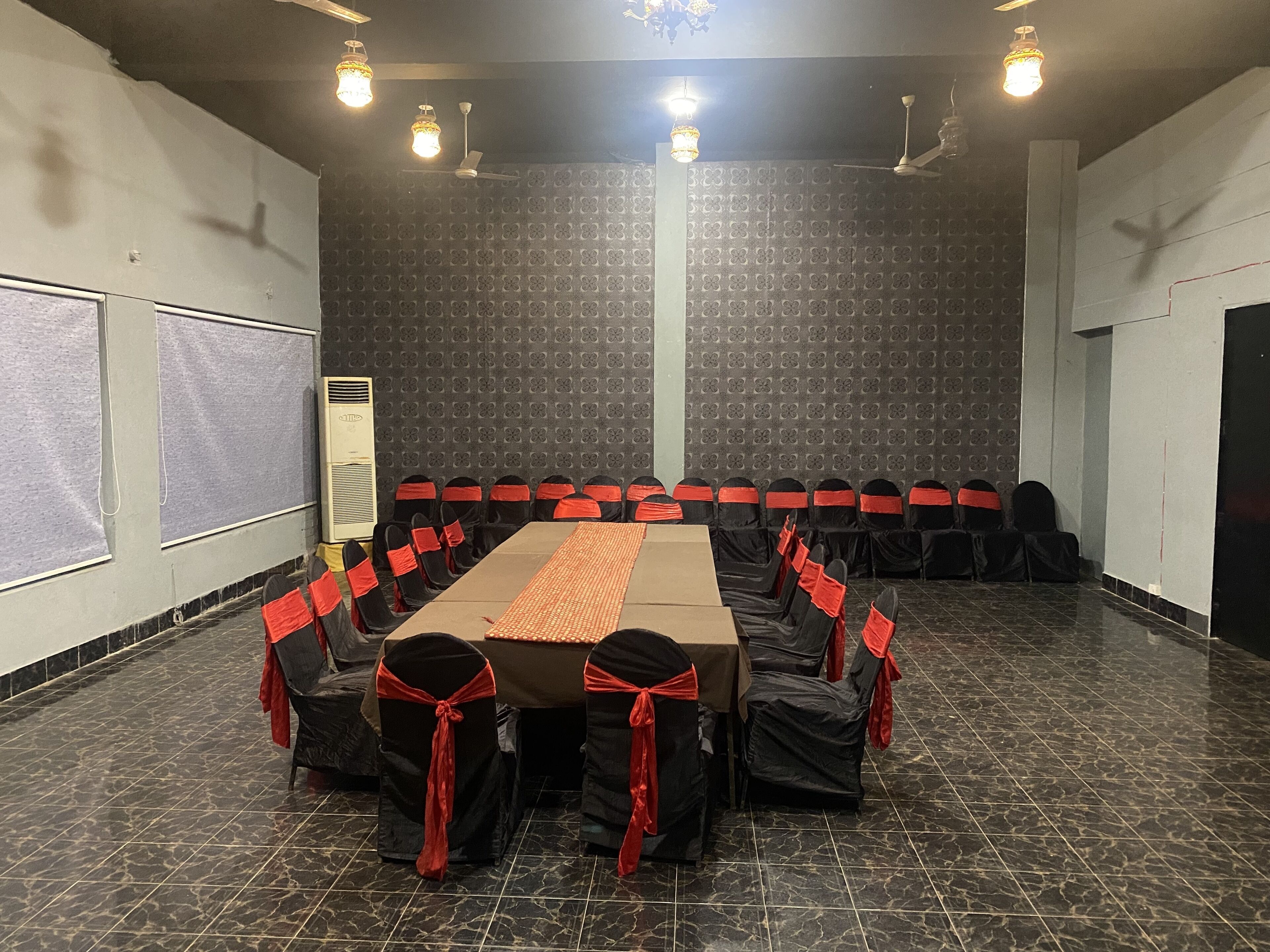 A conference room with a long table, black chairs with red sashes, patterned walls, and ceiling lights.