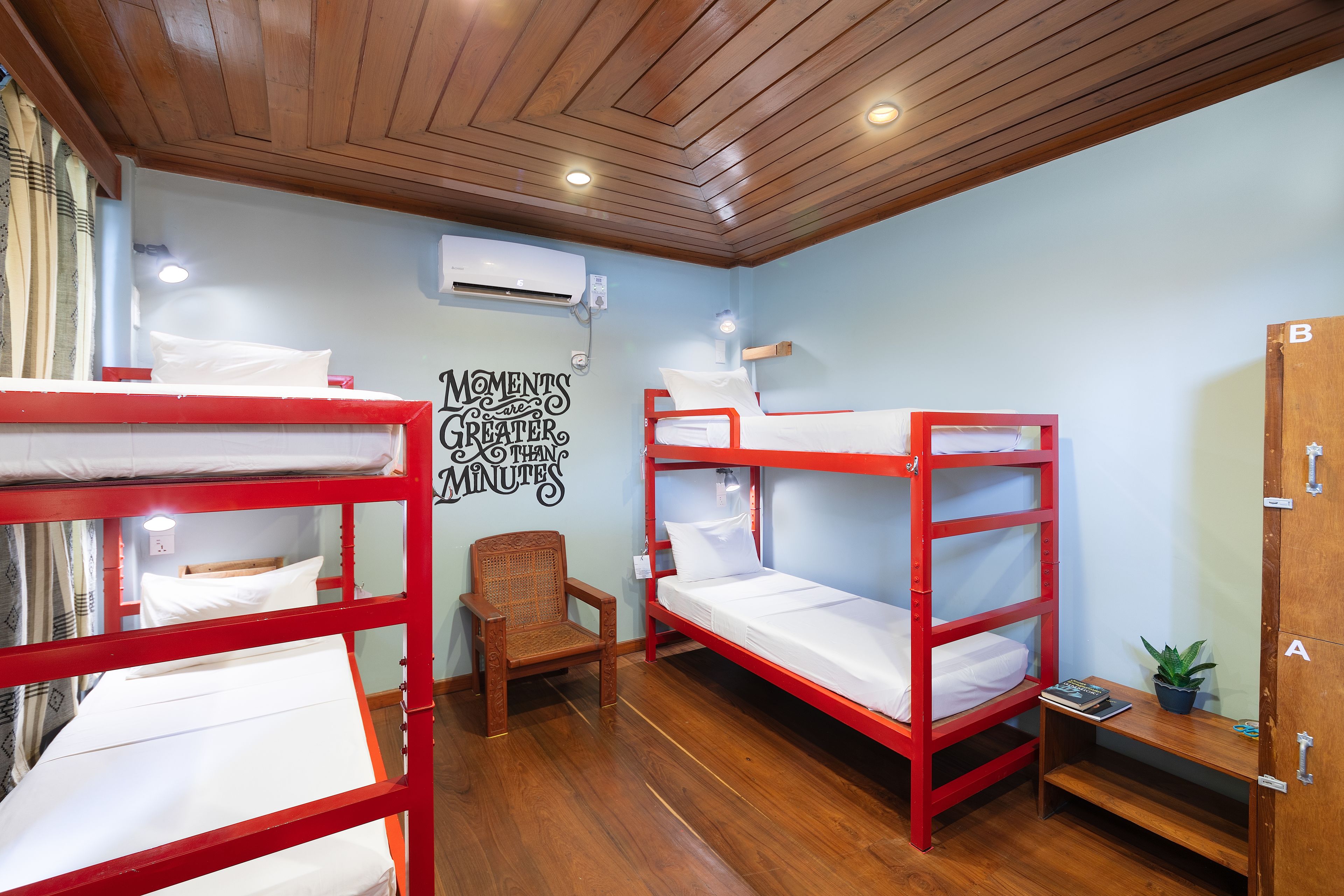 A dormitory room with bunk beds, a wooden chair, a small table with a plant, and a wall-mounted air conditioner.