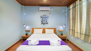 Habitación de hotel con techo de madera, una cama con ropa blanca, un edredón púrpura, dos mesitas de noche con lámparas y un aire acondicionado instalado en la pared.